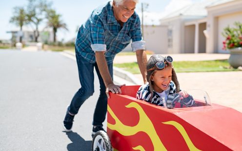 Senior grandfather helping grandson ride on kids toy car. Happy old man pushing car while boy driving on street. Grandparent playing with little boy on gokart.